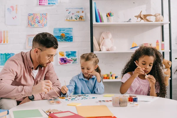 Вчителька в окулярах і багаторасові діти малюють картини з фарбами за столом в класі — стокове фото