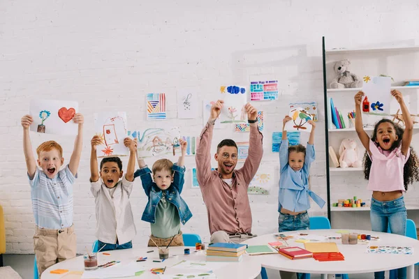 Retrato de professor sorridente e crianças multiétnicas mostrando fotos em mãos em sala de aula — Fotografia de Stock