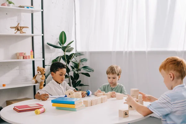 Niños pequeños multiculturales jugando con bloques de madera en la mesa en el aula - foto de stock
