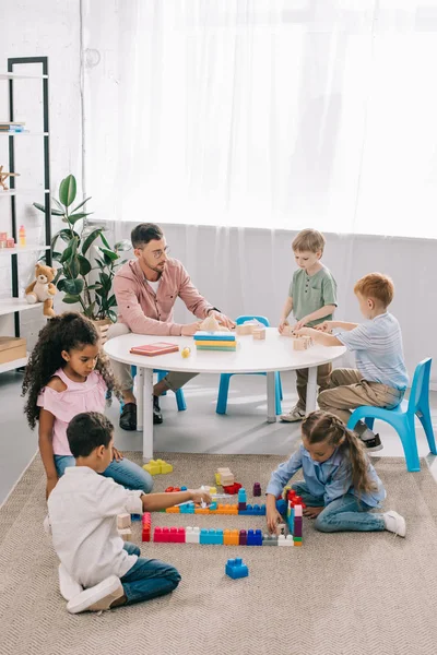 Maestro y niños preescolares multiculturales en el piso con ladrillos de colores en el aula - foto de stock