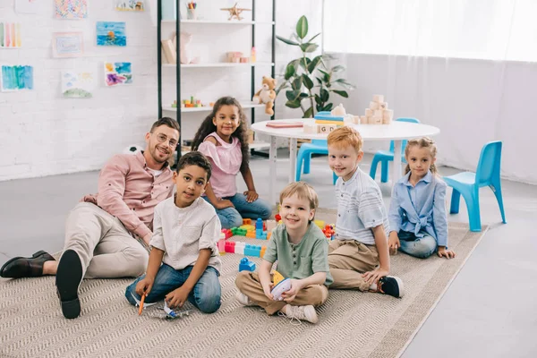Profesor sonriente y niños preescolares multiculturales sentados en el piso con ladrillos de colores en el aula - foto de stock