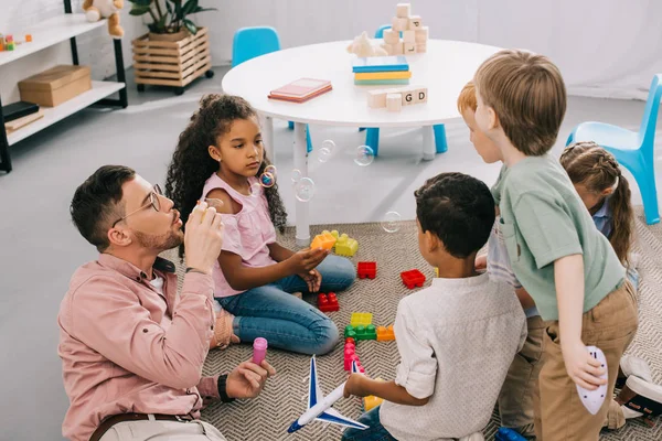 Teacher with soap bubbles and multicultural preschoolers sitting on floor with colorful bricks in classroom — Stock Photo