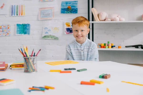 Portrait of smiling red hair boy sitting at table with colorful plasticine for sculpturing in room — Stock Photo