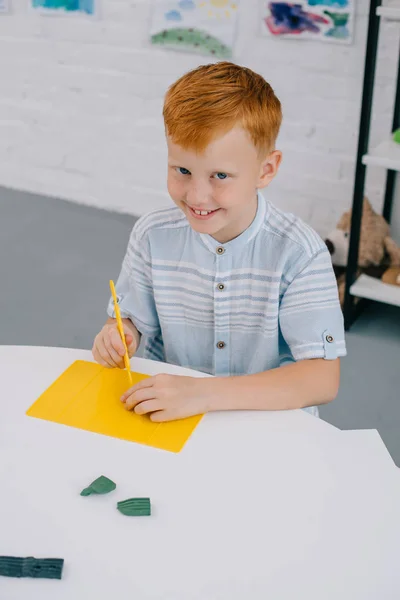 Vue grand angle de sourire garçon aux cheveux roux assis à la table avec plasticine colorée pour sculpter dans la chambre — Photo de stock