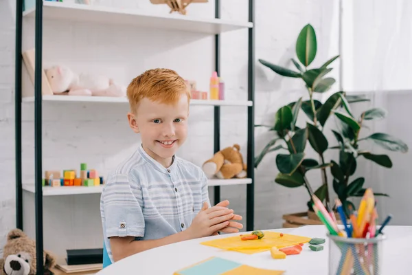 Retrato de niño sonriente pelo rojo sentado en la mesa con plastilina de colores para la escultura en la habitación - foto de stock