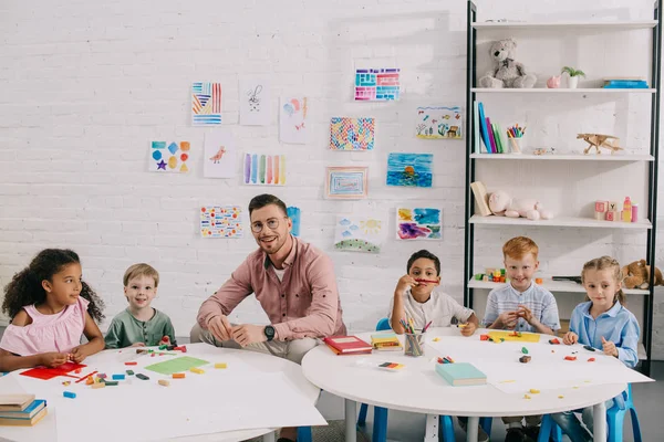 Professeur souriant et enfants multiethniques assis à table avec plasticine colorée dans la salle de classe — Photo de stock