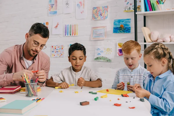 Multiracial preschoolers and teacher with plasticine sculpturing figures at table in classroom — Stock Photo