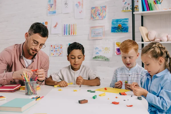 Bambini in età prescolare multietnica e insegnante con figure scultoree in plastilina a tavola in aula — Foto stock