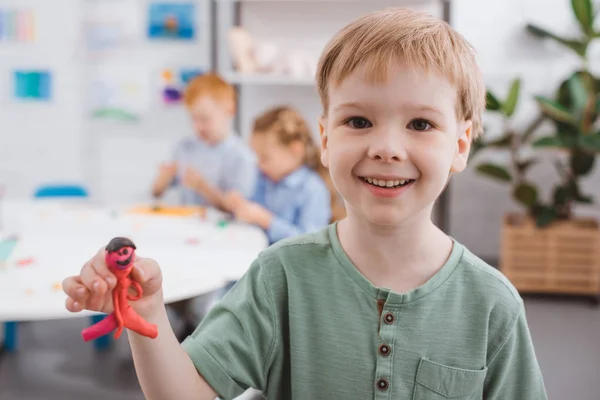 Selective focus of happy kid showing plasticine figure in hand with classmates behind in classroom — Stock Photo