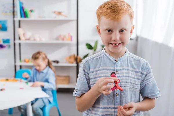 Foyer sélectif de sourire garçon aux cheveux roux avec figure en plasticine et camarade de classe dessin image à la table en classe — Photo de stock