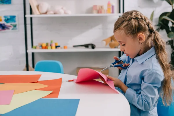 Seitenansicht fokussiertes Kind schneidet Papier mit der Schere am Tisch — Stockfoto