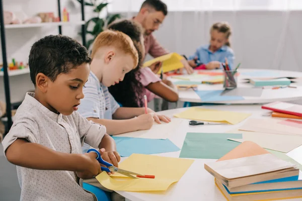 Enfoque selectivo de los maestros y preescolares interracial cortar papeles de colores con tijeras en el aula - foto de stock