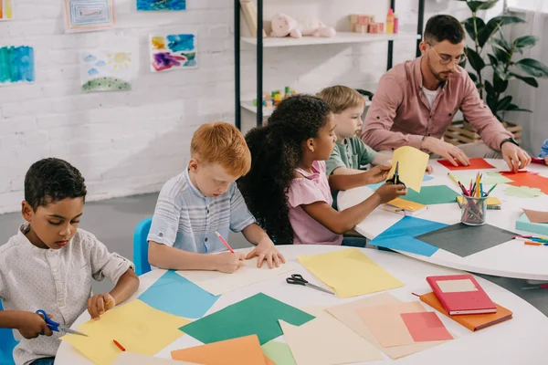 Teacher and multicultural preschoolers cutting colorful papers with scissors in classroom — Stock Photo