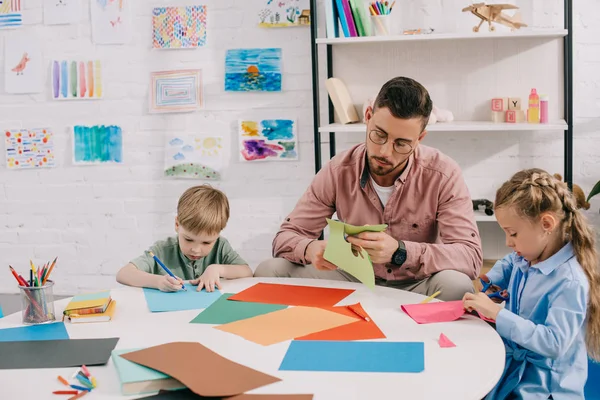 Retrato del maestro enfocado y niños en edad preescolar cortando papeles coloridos con tijeras en el aula - foto de stock