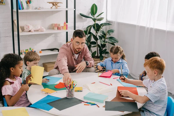 Male teacher and multiracial preschoolers sitting at table with colorful papers in classroom — Stock Photo