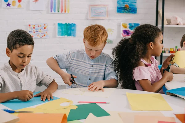 Retrato de niños multiétnicos enfocados con tijeras haciendo apliques de papel en el aula - foto de stock