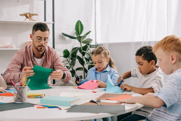 Teacher and multiracial preschoolers cutting colorful papers with scissors in classroom — Stock Photo