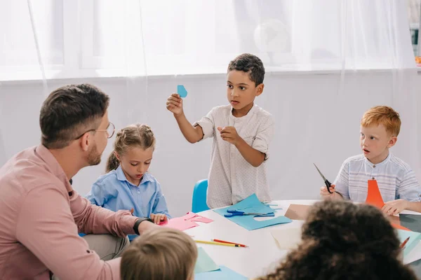 Vista parcial del maestro masculino y los niños preescolares multirraciales sentados en la mesa con papeles coloridos en el aula - foto de stock