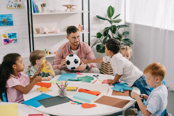 Männlicher Lehrer mit Fußballball und multiethnische Vorschulkinder, die mit bunten Papieren im Klassenzimmer am Tisch sitzen — Stockfoto