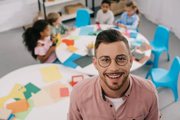 Selective focus of smiling teacher in eyeglasses and interracial kids at table in classroom — Stock Photo