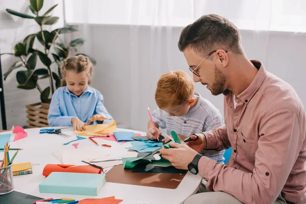 Profesor sonriente y niños en edad preescolar con papeles coloridos y tijeras haciendo apliques de papel en el aula - foto de stock