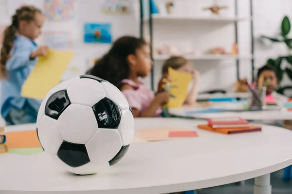 Enfoque selectivo de la pelota de fútbol en la mesa y los niños preescolares multirraciales en el aula - foto de stock