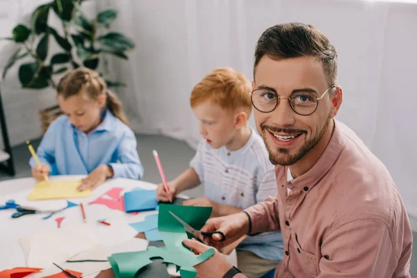 Messa a fuoco selettiva di insegnante sorridente e bambini in età prescolare che fanno applicazione di carta in classe — Foto stock