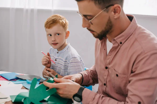 Red hair preschooler looking at teacher cutting paper with scissors in classroom — Stock Photo