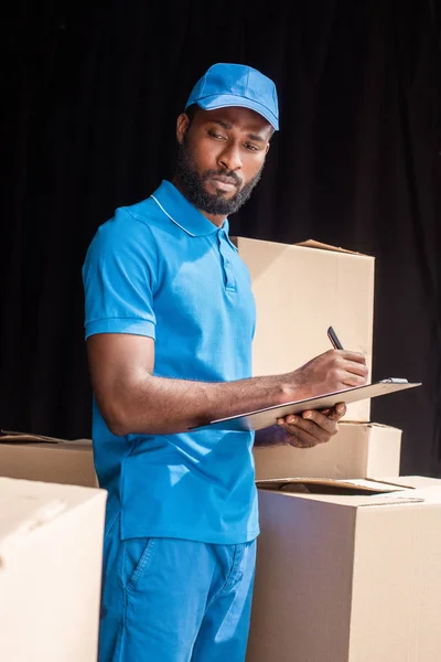 Africano americano repartidor escribir algo a portapapeles y mirando cajas - foto de stock