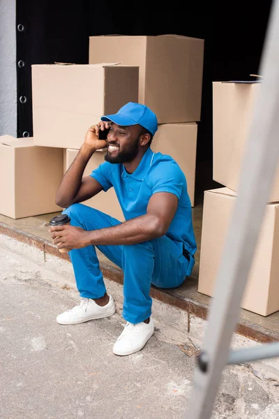 African american delivery man talking by smartphone and holding coffee in paper cup — Stock Photo