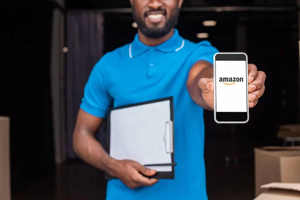 Imagen recortada del repartidor afroamericano mostrando smartphone con página amazónica cargada - foto de stock