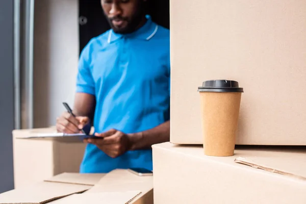 Uomo di consegna afroamericano scrivendo qualcosa negli appunti con il caffè per andare in primo piano — Foto stock