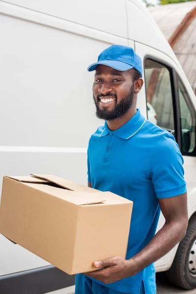 Sonriente africano americano repartidor hombre llevando caja de cartón - foto de stock