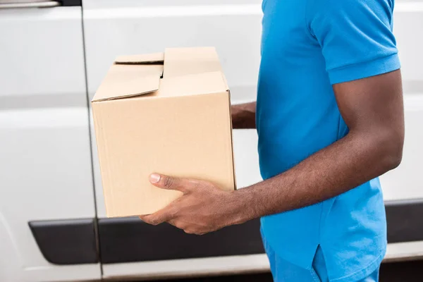 Imagen recortada africano americano repartidor hombre llevando caja de cartón - foto de stock