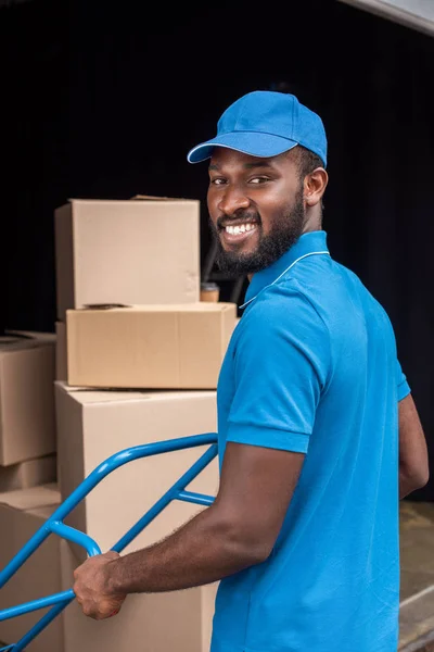 Sonriente afroamericano mensajero con carrito mirando a la cámara - foto de stock