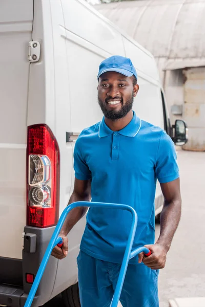 Smiling african american courier with cart looking at camera near van — Stock Photo