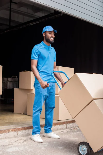 Souriant afro-américain livreur tenant chariot avec des boîtes et détournant les yeux — Photo de stock