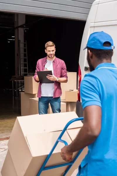 Uomo di consegna africano americano con carrello e scatole a piedi al cliente — Foto stock