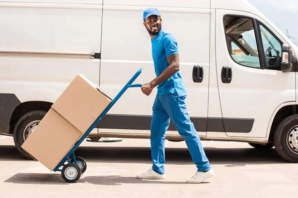 Vista lateral del repartidor afroamericano con carro y cajas - foto de stock