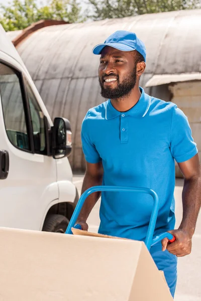 Afro-américain livreur avec chariot et boîtes détournant les yeux — Photo de stock