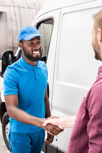 Mensajero multicultural y el cliente estrechando las manos cerca de van - foto de stock