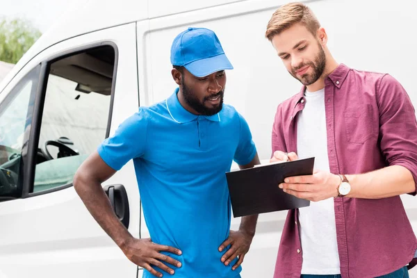 Multicultural courier and client looking at clipboard near van — Stock Photo