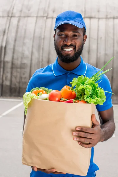 Sonriente africano americano repartidor hombre sosteniendo bolsa de papel con verduras - foto de stock