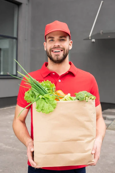 Sonriente caucásico entrega hombre sosteniendo bolsa de papel con verduras - foto de stock