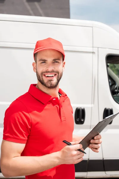 Fröhlicher Zusteller in roter Uniform mit Notizblock, der in die Kamera blickt — Stockfoto