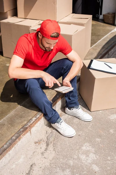 High angle view of young delivery man making calculations on calculator near cardboard boxes — Stock Photo