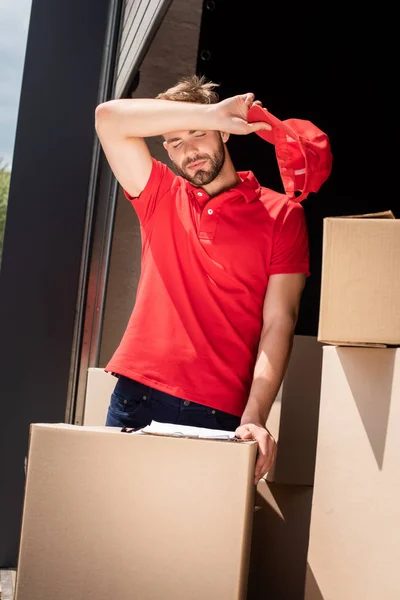 Portrait of tired delivery man standing near cardboard box with notepad — Stock Photo