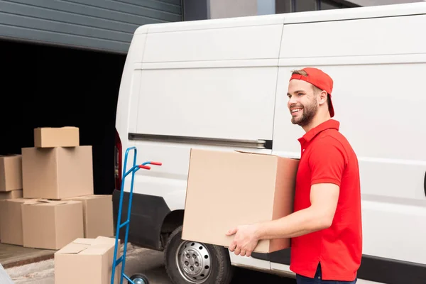 Smiling delivery man in uniform with cardboard box standing near white van in street — Stock Photo