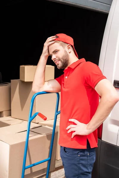 Young delivery man in red uniform  leaning on delivery cart — Stock Photo