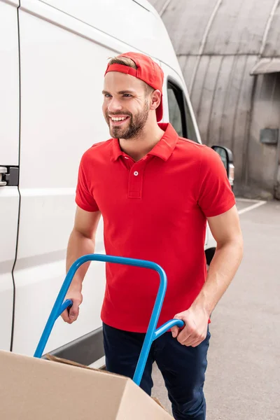 Homem de entrega sorridente com caixa de papelão no carrinho de entrega — Fotografia de Stock